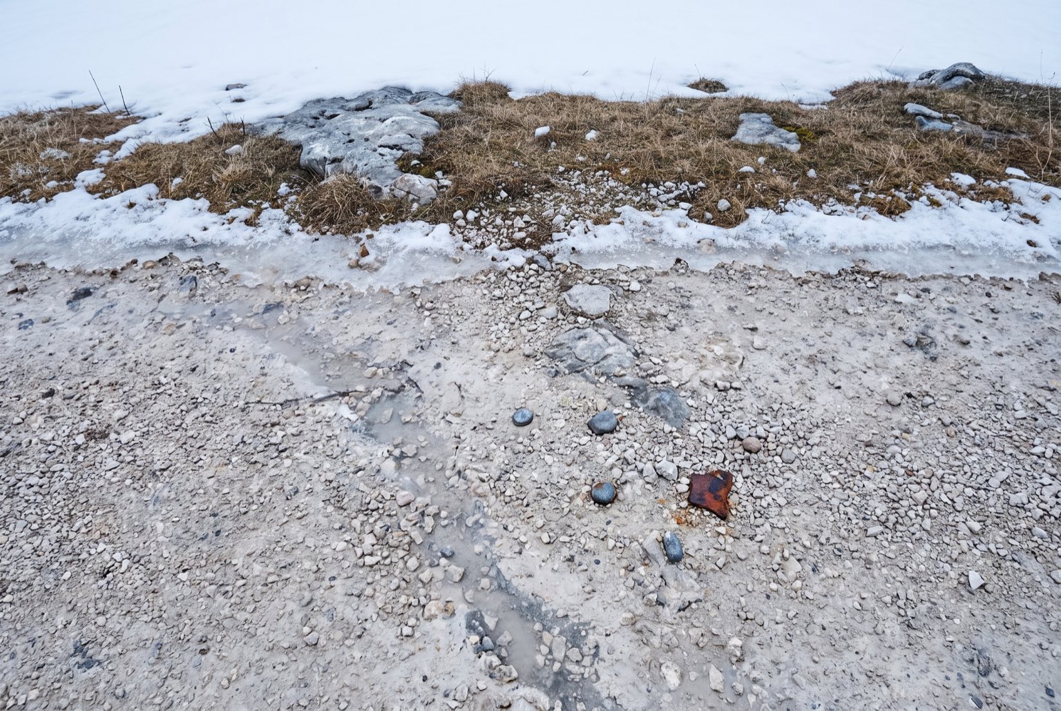 Close overhead view of a coarse pale limestone gravel road surface, wet from melting snow, with old snow at the verge.