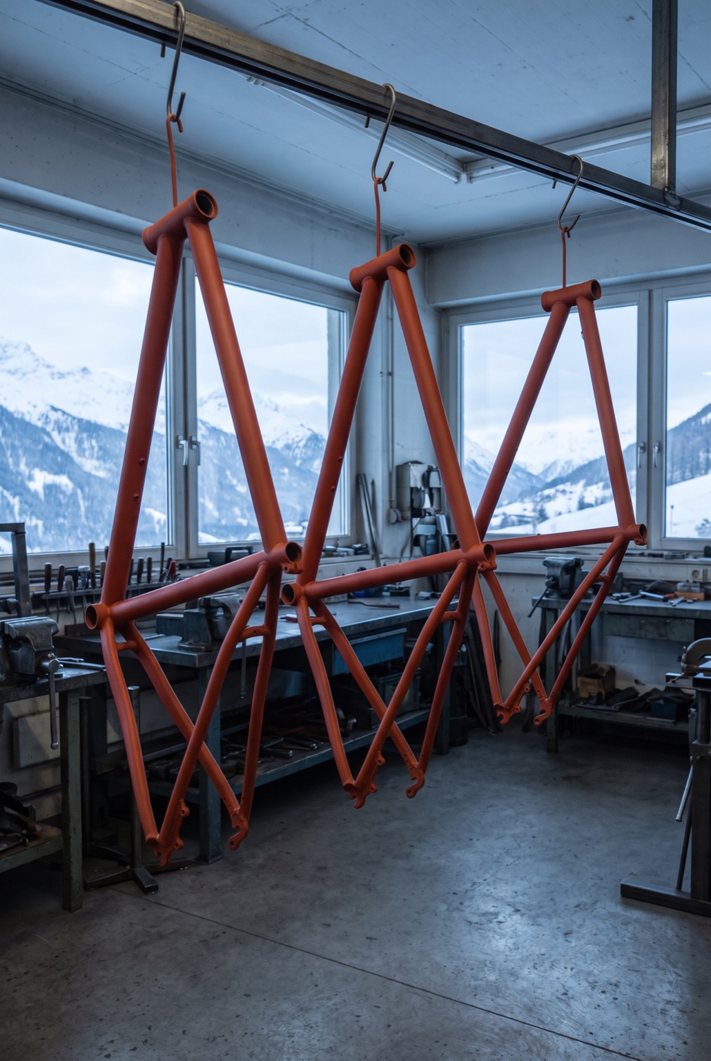 Three bare steel bicycle frames in matte orange-red lead-oxide primer hanging by their head tubes from a rail in a bright workshop.