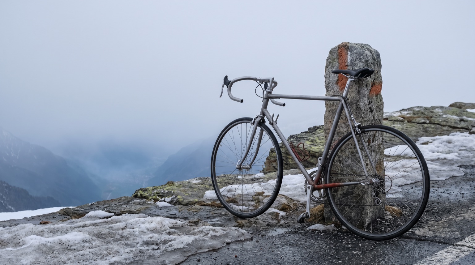 A plain steel road bicycle leaned against a stone snow-marker post at the top of an alpine pass, patches of old snow, the valley falling away into cloud behind.